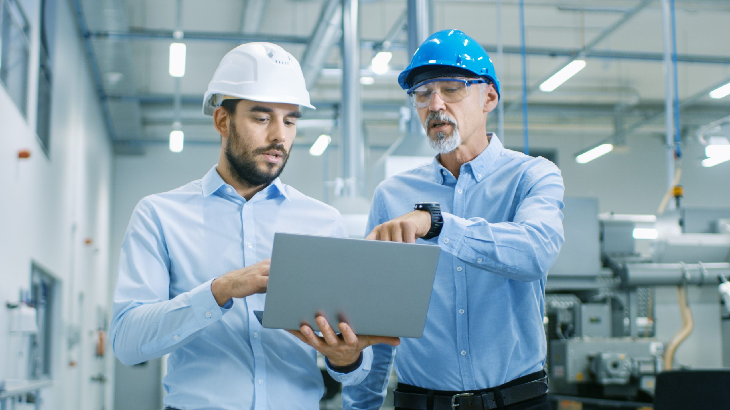 worker with glasses explaining work to his co-worker with white helmet through laptop